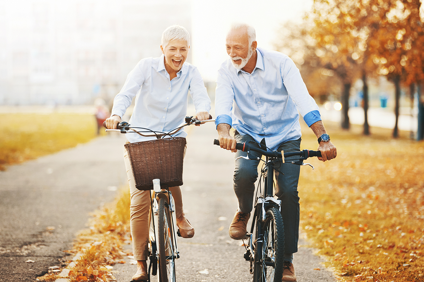 Closeup front view of couple of cheerful seniors enjoying a bicycle ride in the city streets. It's mild October day with orange leaves and grass around them.
